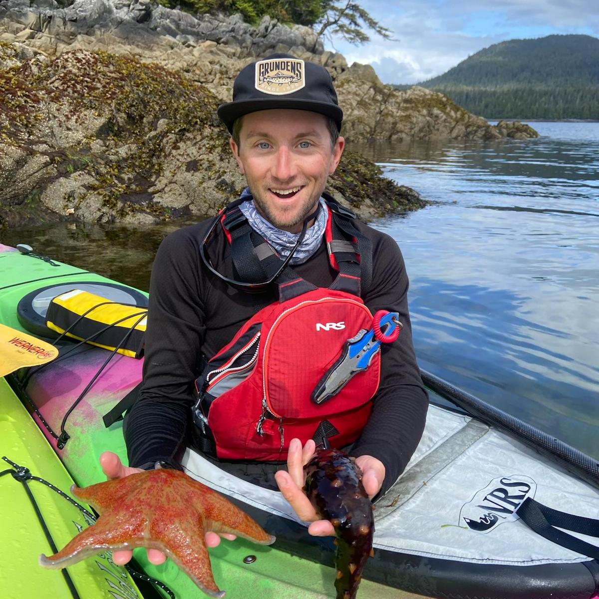 Person in kayak holding a starfish and sea cucumber, surrounded by rocky shore and forest.